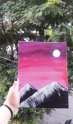 someone is holding up a painting with mountains in the background and a full moon above them