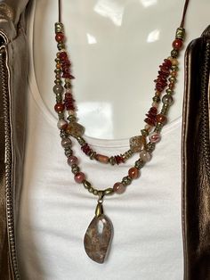 A 1 1/4 inch Peanut Obsidian stone dangles from a strand of Noreena Jasper, Carnelian and Bronze beads on 1mm brown leather while another strand of Amber, Brown and Green Opal, Carnelian and Bronze beads hangs above. Around the neck is 20" and total length of necklace is 24" all strung on two 1mm strands of bronze leather with a Bronze toggle to close. No lead or nickel used. Please use the Last On, First Off approach. Stones discolor with lotions, perfumes, hairspray, sweat, chlorine, even some Bohemian Gemstone Beads And Cabochons In Brown, Bohemian Brown Gemstone Beads And Cabochons, Bohemian Brown Gemstone Beads, Noreena Jasper, Amber Brown, Obsidian Stone, Beaded Statement Necklace, Green Opal, Stone Pendant