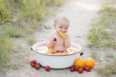 a baby sitting in a tub with strawberries and oranges on the ground next to it