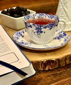 an open book on a wooden table next to a cup and saucer with berries in it