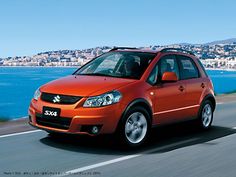 an orange small car driving down the road near water and buildings in the background with blue sky