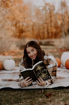 a woman laying on top of a blanket holding an open book in her hands and smiling at the camera