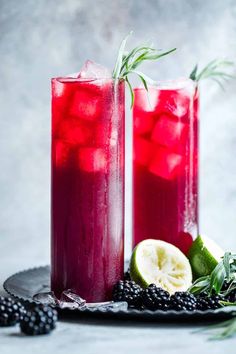 two glasses filled with red liquid and garnish next to blackberries on a plate