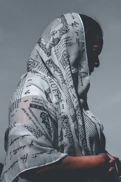 a woman wearing a white and black robe with writing all over her face, standing in front of a cloudy sky