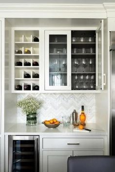 a kitchen with white cabinets and glass shelves filled with wine glasses, oranges and bottles