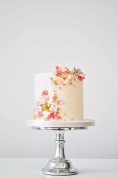 a white cake with pink and yellow flowers sitting on top of a metal stand in front of a white wall