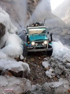 a green truck driving through some ice covered rocks and water with mountains in the background