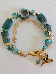 Lovely double strand beaded Premium Czech glass flower bracelet featuring table cut matte aqua with bronze rectangular beads, gold etched ivory/aqua rondelles, fire polished aqua Aurora Borealis crystals, and single 14 mm turquoise Picasso pansy flower. All accented with antique gold plated daisies and shiny gold cubes. Satin Hamilton rose chain adds the perfect delicate second strand. Premium gold electroplated brass chain in satin finish with intricate rose flower pattern. Bracelet is beaded o Rectangular Gold Jewelry With Gemstone Beads, Adjustable Gold Beaded Bracelets Made Of Czech Glass, Adjustable Gold Beaded Bracelets With Czech Glass, Gold Rectangular Jewelry With Gemstone Beads, Gold Bracelets With Faceted Beads, Unique Gold Beaded Bracelets Nickel Free, Elegant Gold Bracelets With Czech Glass, Blue Double Strand Beaded Bracelets As Gifts, Adjustable Gold Beaded Bracelets In Czech Glass