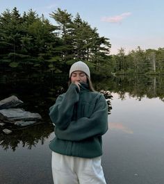 a woman standing in front of a body of water wearing a green jacket and white pants