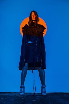 a woman standing in front of a blue background with an orange circle behind her and the top half of her coat pulled back