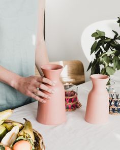 a woman holding two pink vases on top of a table next to fruit and vegetables