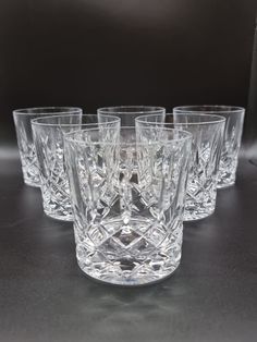 six crystal tumblers are lined up on a black table top, with one empty glass in the middle