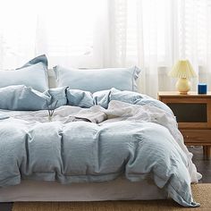 an unmade bed with blue linens and pillows in a white room next to a window