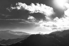 black and white photograph of the sun shining through clouds over mountain tops with trees in foreground