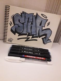 a notebook with graffiti written on it next to four markers and a marker pen in the foreground