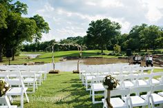 an outdoor ceremony setup with white chairs and flowers on the aisle, in front of a pond