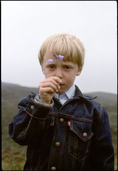 a young boy holding something up to his mouth while standing in front of a field