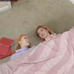 two young women laying on the floor under a pink and white striped comforter, looking at each other