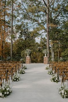 an outdoor ceremony setup with wooden chairs and white flowers on the aisle, surrounded by tall trees