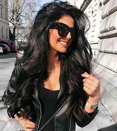 a woman with long black hair and sunglasses standing next to a wall wearing a leather jacket