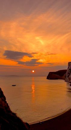 the sun is setting over the ocean with rocks in the foreground and people walking on the shore