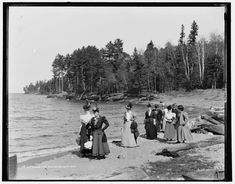 an old black and white photo of people standing on the shore of a body of water