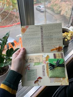 someone holding up an open book in front of a window with plants and flowers on it