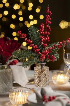 the table is set for christmas dinner with red berries and greenery in glass vases