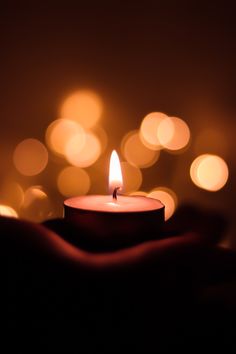 a lit candle in someones hand with blurry lights behind it on a dark background