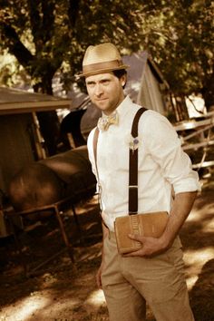 a man wearing a hat and suspenders is standing in the shade with his hands on his hips