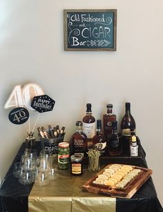 an assortment of liquors and snacks on a table with a chalkboard sign above it