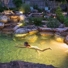 a person swimming in a pool surrounded by rocks and plants at night with lights on