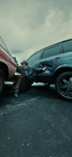 two people sitting on the ground next to a parked car and another person leaning over