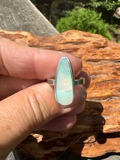 Another dreamy opal from Lightning Ridge.this one is a unique  light green color with pink and gold shimmers streaking through the middle. So lovely!  I got this beauty as a rub so I was able to shape, polish and set her in a fine and sterling silver setting in a flat wire band.  The stone is 21 mm x 8 mm (approximately .83 inches x .4 inches). Boulder opals are mined from ironstone boulders under the ground.  The opal is formed from a solution of silicon dioxide and water.  As the water runs through the earth, it picks up silica from sandstone a day carries this silica-rich solution into cracks and voids, caused by natural faults or decomposing fossils.  As the water evaporates, it leaves behind a silica deposit, which can be found in one or more colors, like blue, purple, red, green, yel Iridescent Opal Ring With Gemstone, Unique Handmade Iridescent Opal Ring, Unique Oval Opal Ring For Healing, Green Opal Cabochon Ring, Iridescent Oval Opal Ring, Unique Iridescent Opal Ring, Unique Green Opal Ring, Boulder Opal Ring, Flat Wire