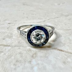 Breathtaking handcrafted platinum Art Deco style diamond and sapphire halo engagement ring. The center stone is a lovely bezel-set 0.50 carat old European cut diamond. The stone is G-H color VS2 clarity. It is haloed by a row of fine French cut sapphires weighing approximately 0.45 carat. The ring is accented with 4 round diamonds on the shoulders, weighing approximately 0.04 carat. G-H VS. Fine milgrain adds to the delicacy of the ring. Beautiful openwork filigree decorate the under gallery. Di Art Deco Diamond Ring With Bezel Setting, Classic Sapphire Diamond Ring With Bezel Setting, Classic Round Sapphire Ring With Bezel Setting, Heirloom Sapphire Halo Ring, Heirloom Sapphire Diamond Ring With Bezel Setting, Anniversary Halo Ring With Lab-created Sapphire, Sapphire Diamond Ring With Rose Cut Diamonds, Art Deco Sapphire Ring With Bezel Setting, Classic Platinum Sapphire Ring With Bezel Setting