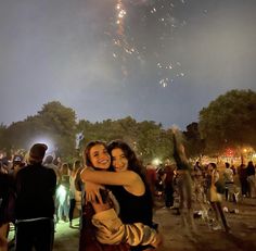 two women hugging each other while fireworks are in the sky above them and people standing around