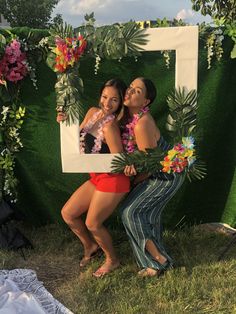 two women pose for a photo in front of a backdrop with flowers and plants on it