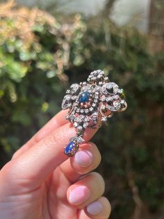 A literal bouquet of jewels! This stunning Sapphire and Diamond brooch is a feast for the eyes, composed in a design reminiscent of Victorian era Giardinetti (little gardens) jewelry. Crafted in antique 14 carat gold, this brooch features a dazzling array of 78 miniature bezel-set diamonds, arranged in a botanical motif. Dotted along the corners of the brooch are 7 round mix cut emeralds, which add a whimsical flair to the silhouette. The centerpieces of this adornment however are the two vibrant blue cabochon-cut sapphires: the first is nestled in the very center of the brooch, surrounded by a halo of diamonds, while the second dangles at the bottom like an elegant flower bulb. A perfect mix of glamour and lushness, this treasure is the perfect addition to any antique jewelry collection! Antique Baroque Brooches With 17 Jewels, Ornate Baroque Jewelry With Rose Cut Diamonds, Elegant Diamond Multi-stone Brooch, Ornate Rose Cut Diamond Brooches For Weddings, Ornate Diamond Brooch For Anniversary, Ornate Diamond Brooches For Anniversary, Antique Diamond Brooch Jewelry, Victorian Diamond Brooch For Evening, Antique Multi-stone Cluster Jewelry