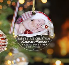 a baby's first christmas ornament hanging from a christmas tree with lights in the background