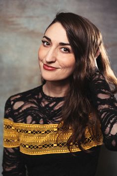 a woman with long hair wearing a black and yellow top smiles at the camera while standing in front of a gray background