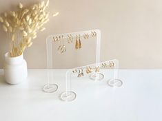 three clear acrylic jewelry stands on a white table next to a vase with dried flowers