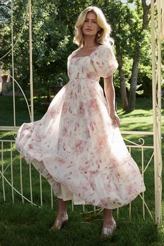 a woman in a dress standing on the grass near a gazebo with trees and bushes behind her