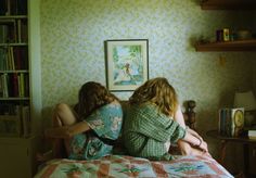 two women sitting on top of a bed in a bedroom next to a book shelf