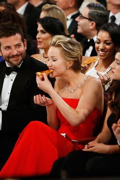 a man and woman sitting next to each other in front of an audience eating pizza