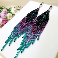 purple and blue seed beaded earrings sitting on top of a white table next to flowers