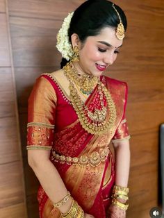 a woman in a red and gold sari with jewelry on her neck, standing next to a wooden wall