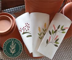 three white towels with red and yellow flowers on them sitting next to potted plants