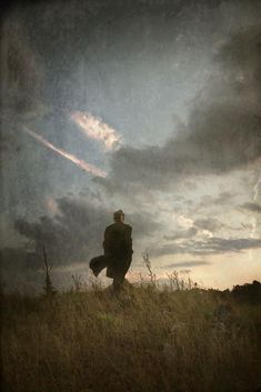 a man standing on top of a grass covered hill under a cloudy sky with clouds