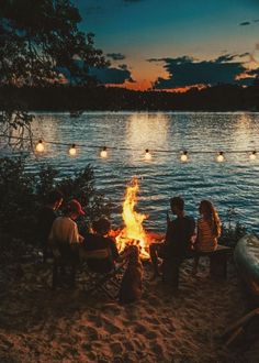 four people sitting around a campfire with lights strung over the water in the background