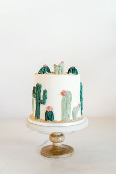 a white and green cake with cactus decorations on the top is sitting on a pedestal